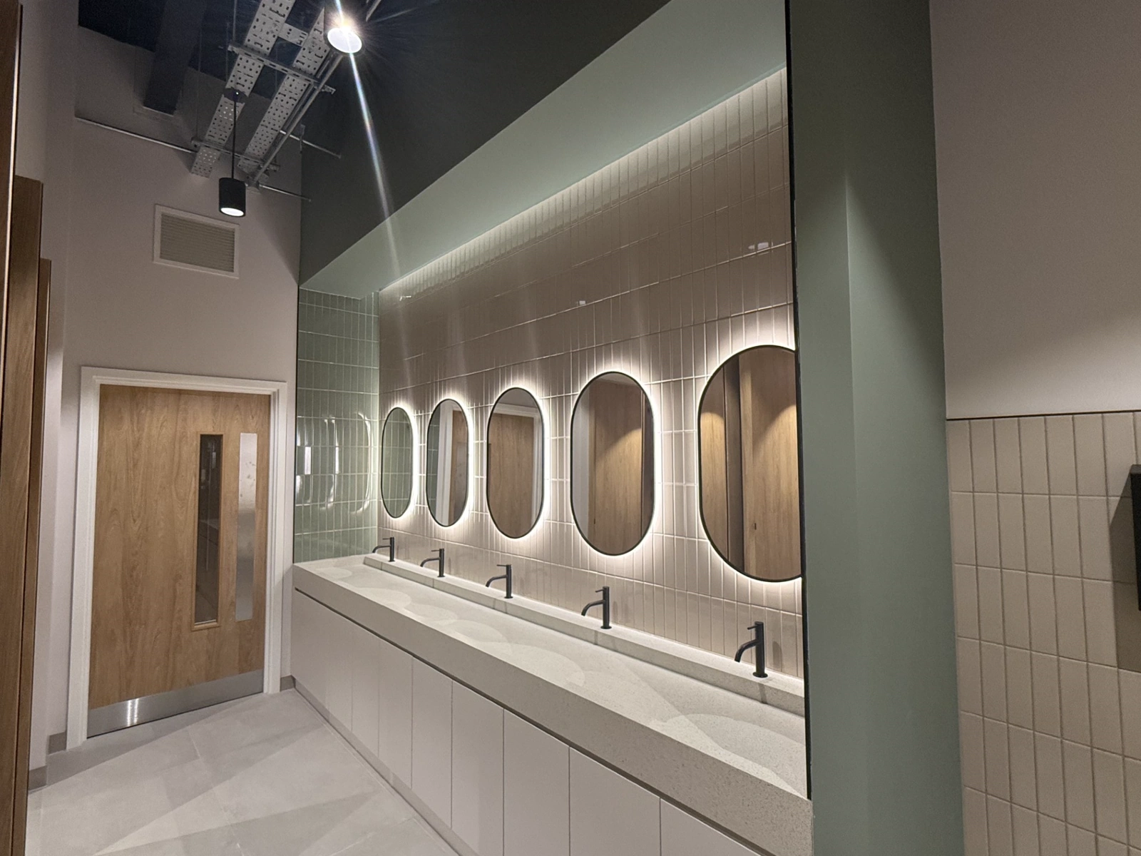 Washroom at Broadgate Quarter with sage green painted walls, tiled surrounds and backlit oval mirrors